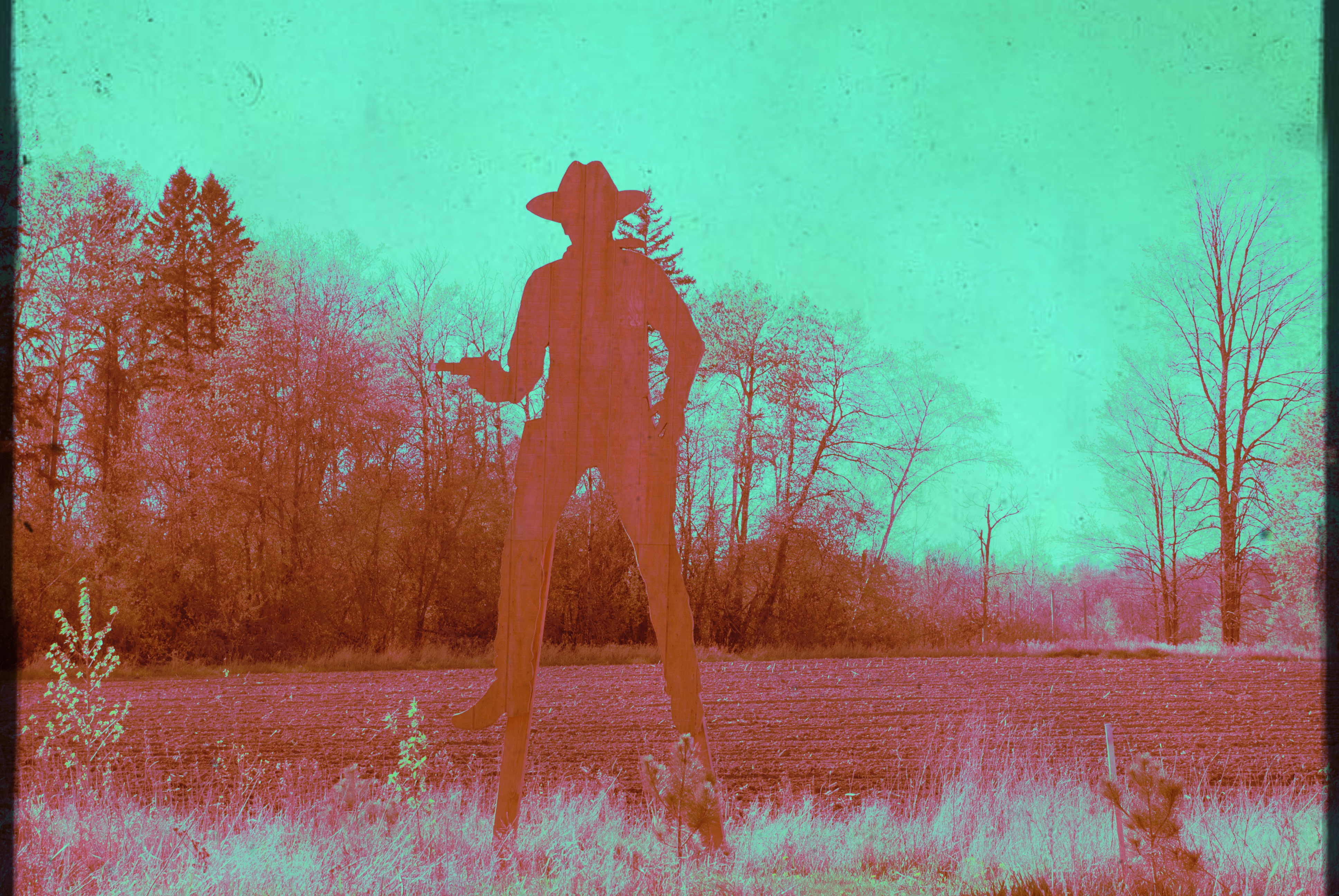 A tall wooden silhouette of a cowboy on stilts, towering over a meadow and plowed field, with a copse of trees in the background. The image has been modified to have unnatural pink and green tones, with a grungy overlay. 