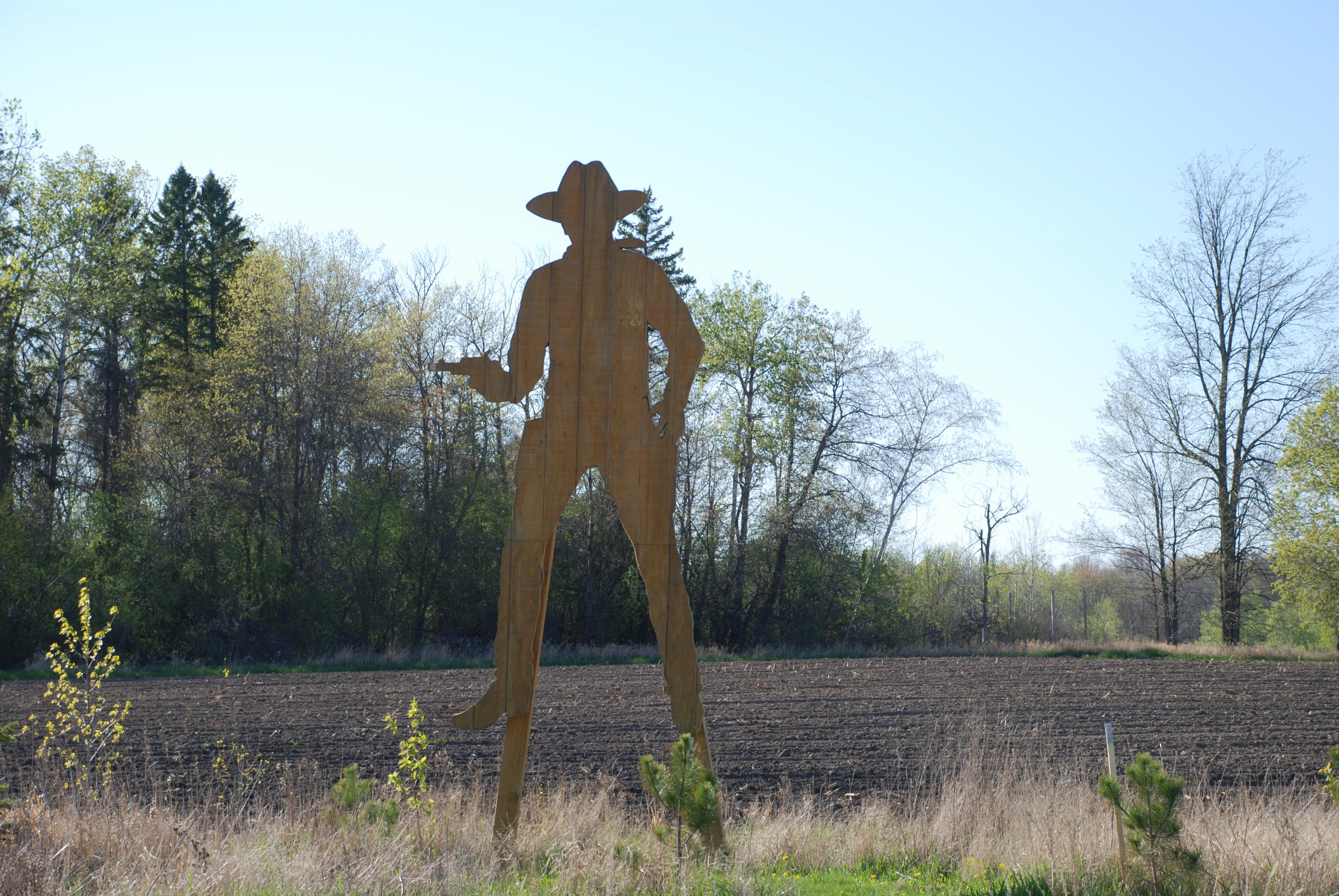 A tall wooden silhouette of a cowboy on stilts, towering over a meadow and plowed field, with a copse of trees in the background.
