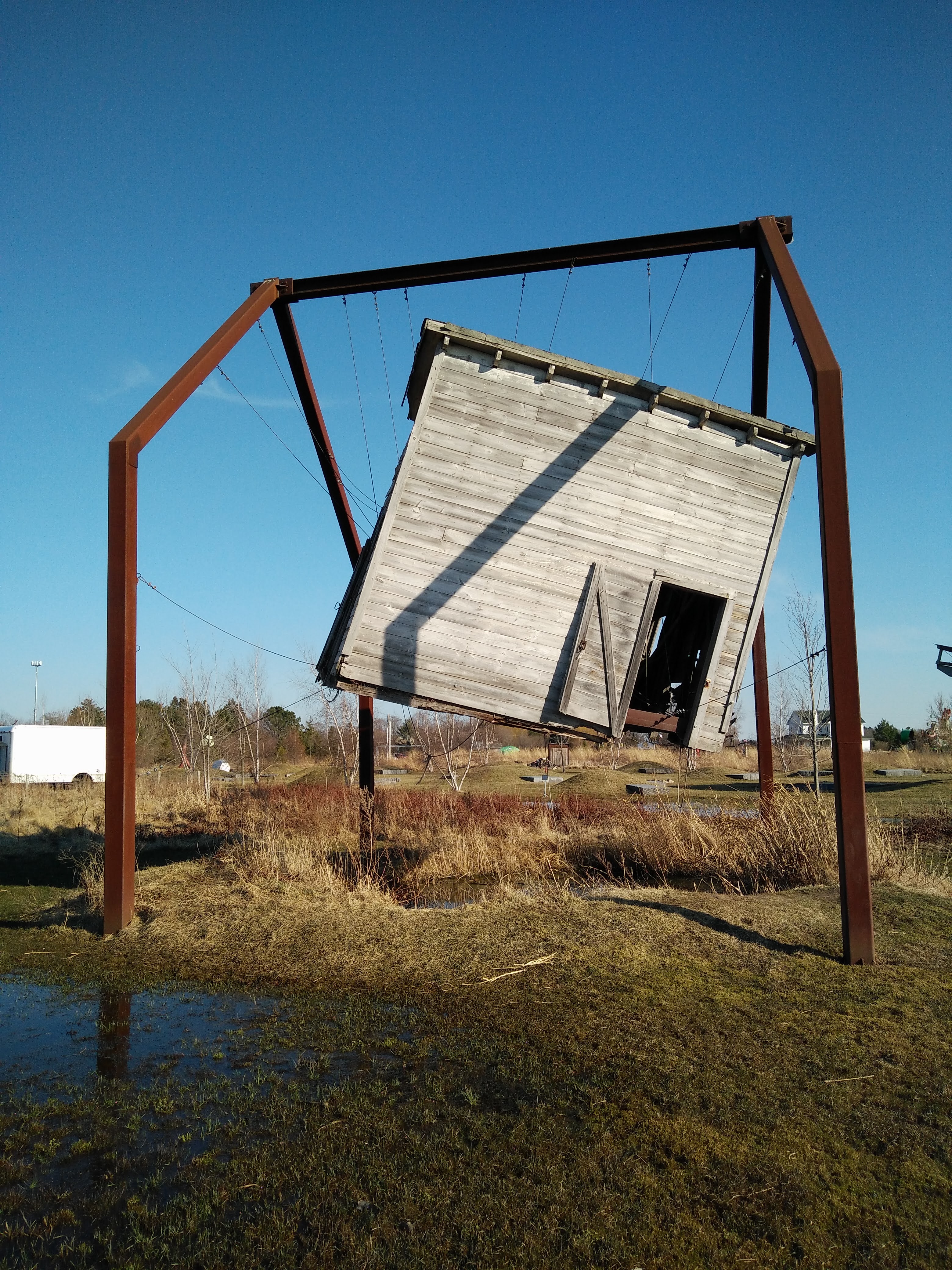 A sculpture of a large, decaying shed, suspended above the ground by a series of ropes woven through the shed and attached to metal beams. 
