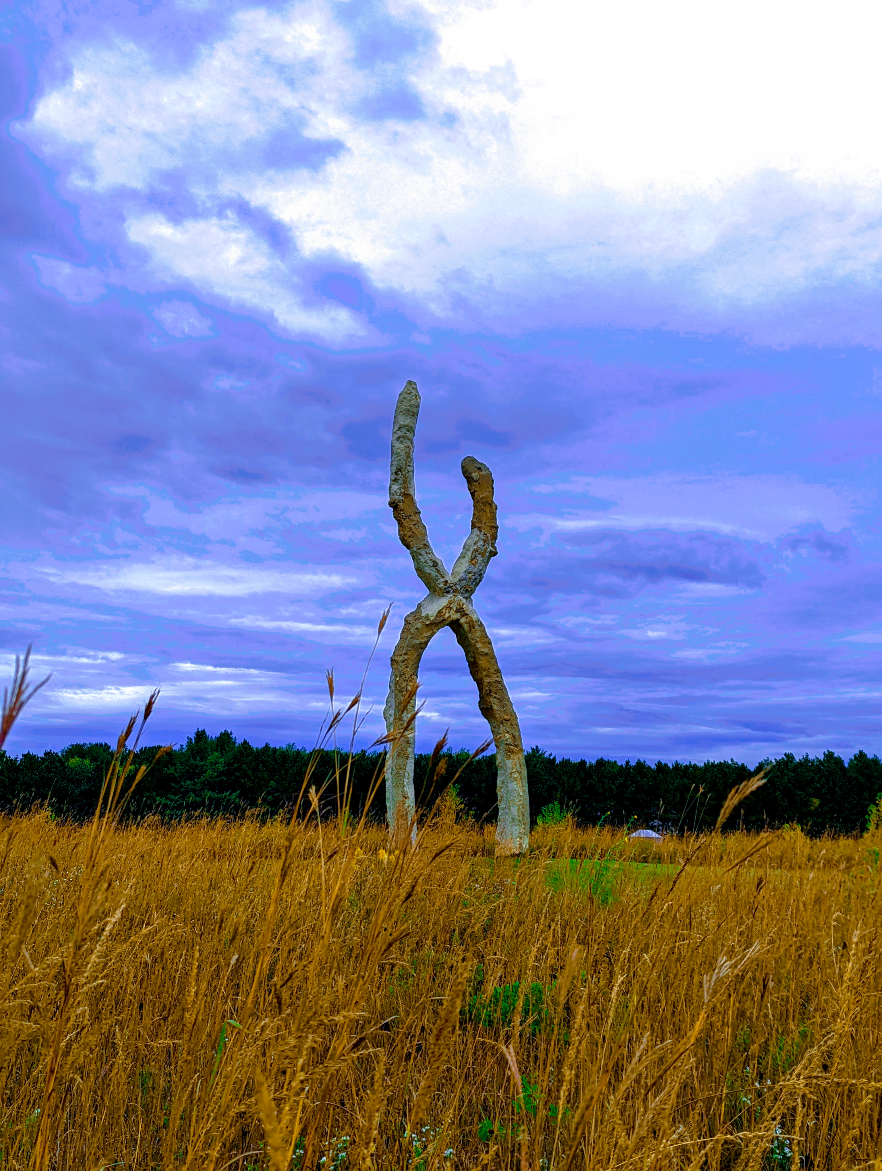 An enormously large concrete, steel and earthen sculpture in the shape of a large X. It stands tall over the prairie landscape and trees. The colours of the photograph have been modified to accent and brighten it, which gives the sky a purplish-blue colour and the colours of the sculpture itself and the golden prairie grasses are also accentuated and stand out compared to the original.