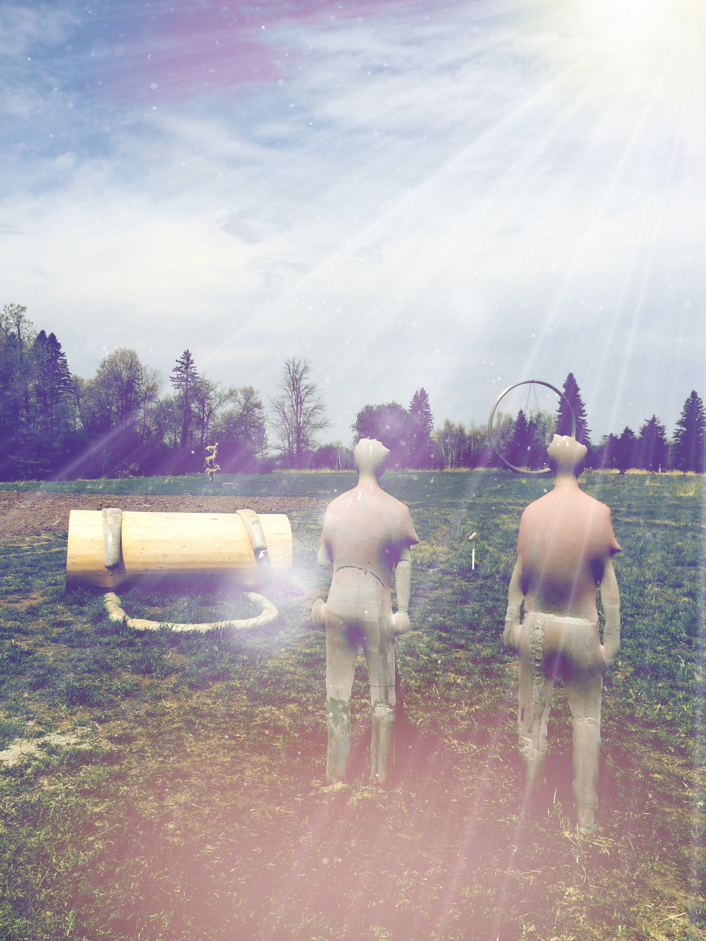 A sculpture composed of two human figures and a large hollow wooden tube. The figures are facing away from the viewer, out across the wider sculpture park. In the distance are two unrelated sculptures, one of a series of taxidermied deer and one of a large round circular structure, and the varied treeline. The original photo has been modified to add a strange and slightly retro feel, adding light auras and rays of light shining down over the scene from the upper right corner of the photograph.