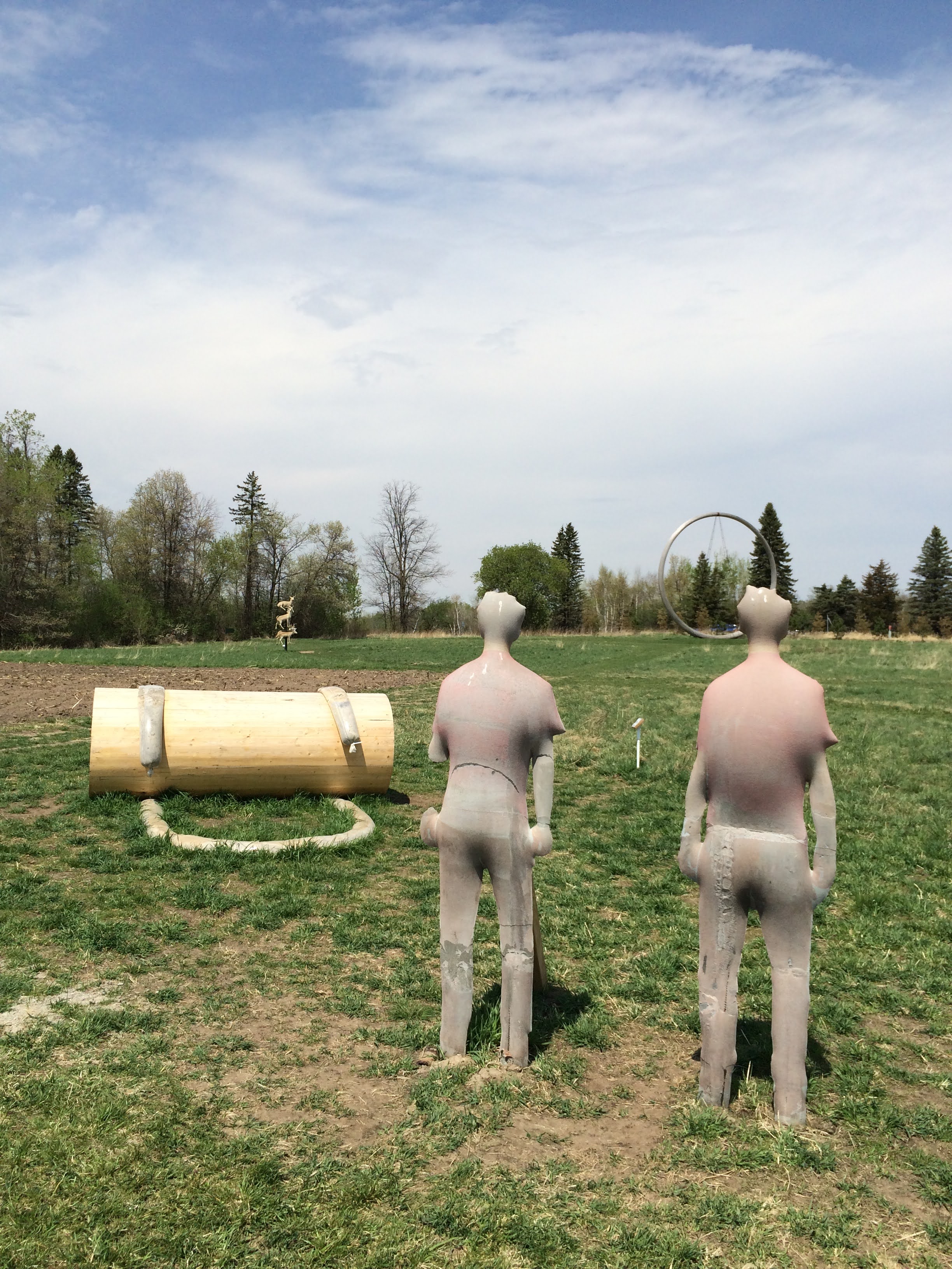 A sculpture composed of two human figures and a large hollow wooden tube. The figures are facing away from the viewer, out across the wider sculpture park. In the distance are two unrelated sculptures, one of a series of taxidermied deer and one of a large round circular structure, and the varied treeline.